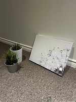 Canvas print with dandelion design and two artificial plants in pots on carpeted floor against wall