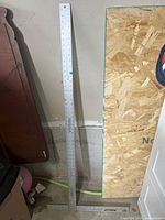 Full view of the metal drywall square standing upright against a wall next to wood and other objects, showing the entire length and markings along the rule edge.