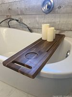 Photo shows wooden bath tray resting on edge of bathtub with three ivory flameless candles on top; tray features recessed cup holder and accessory slot.