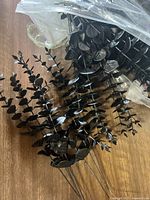 Set of multiple black plastic artificial plant stems with wire bases, shown spread out on a wooden surface under clear plastic wrap.