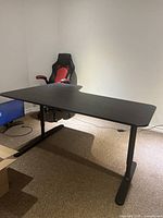 Black right corner office desk with a veneer finish and adjustable height, shown in room corner with office chair and monitor nearby.