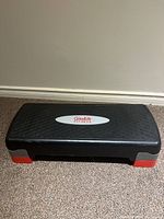 Goodlife Fitness aerobic stepper viewed frontally showing textured surface, logo, and adjustable red and gray feet on a carpeted floor.
