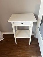 Front view of white IKEA Hemnes nightstand showing drawer and lower shelf, minor scratch visible on top surface.
