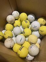 Box full of mixed white and yellow golf balls with multiple brands visible, some with markings and wear showing they have been used.