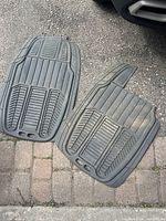 Two Michelin black rubber car floor mats side by side on pavement with textured grooves and securing holes visible.