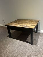Square modern dark wood table with visible natural wood grain on tabletop, black wooden legs, placed on carpeted floor.