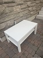 Side view of the white rectangular coffee table showing the lower shelf and four legs, positioned on a brick floor against a stone wall.