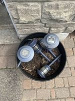 Five blue outdoor solar garden lights placed inside one round black barrel filled with soil on outdoor sidewalk