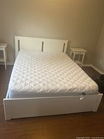 Front view of the white queen-sized bed frame with mattress (mattress not included). Wooden frame with cream white finish on a brown hardwood floor.
