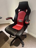 Front and angled view of a red and black faux leather gaming chair showing wear and tear on seat and lumbar area, padded armrests, reclining non-adjustable back, black base with wheels.