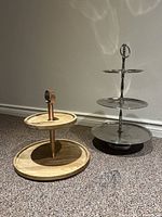 Both serving platters displayed side by side on the floor. One is silver metal with three tiers, the other is wooden with two tiers and a copper handle.