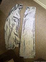 Two silver polyester curtain panels laid out on carpet, showing the metal grommet tops and wrinkled fabric texture.