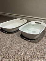 Two white stoneware baking dishes side by side on carpeted floor; one has a glass lid with white handle, the other is uncovered. Both have integrated handles.