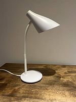 White plastic desk lamp with a conical shade and round base on a wooden surface, showing the full lamp upright with the neck slightly bent.