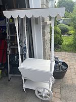 Full view of the white wooden decorative display cart with canopy featuring scalloped design and wheels, positioned on a floor near outdoor garden area.