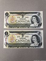 Two 1 dollar Canadian bank notes, one above the other, displayed flat on a surface showing front side with Queen Elizabeth II portrait and Canadian coat of arms.