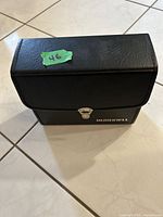 Black Bushnell carrying case closed, standing on tiled floor, with silver clasp and Bushnell logo.