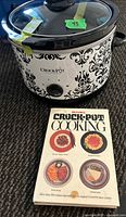 Full view of white Crock Pot slow cooker with black damask pattern and glass lid, alongside Rival Crock-Pot cooking cookbook with recipe images on cover.