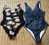 Two one-piece swimsuits laid flat: floral print with black sides on left, solid navy blue on right, showing full front view.
