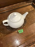 White porcelain teapot with round body and curved spout displayed on a clear ornamental base on a wooden surface.