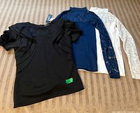 Three designer women's tops displayed together on a surface; includes black short-sleeve top and blue and white long-sleeve lace overlay tops.