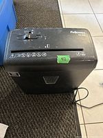 Front view of black Fellowes Powershred H-6C paper shredder on carpeted floor, showing feed slot, control knob and green numbered sticker on top.