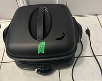 Full view of black Hamilton Beach 38500 electric skillet with lid and attached power cord, placed on tiled floor.