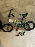 Full side view of a green and white 16-inch kids bike with black tires and black handlebars. A pair of detachable training wheels lies in front of the bike.
