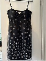 Front view of the black spaghetti strap dress showing the silver and rose gold circular patterns on fabric. Dress is hanging on a black hanger against a white wall.
