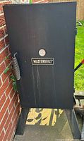 Front view of black Masterbuilt electric smoker, showing the logo and thermometer on the door and spring handle on the left.