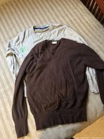 Two men's sweaters laid flat on striped beige surface; brown cashmere V-neck sweater over grey long sleeve crewneck sweater.
