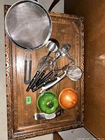 Overall view of the kitchen utensils spread on a wooden table, showing strainers, ladle, peelers, grater, and ceramic bowls.