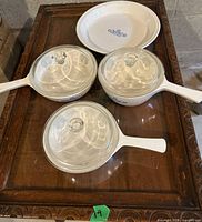Three white Corning Ware saucepans with lids and round pie dish on wooden table