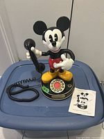 Front view of the Mickey Mouse talking telephone on a blue plastic container lid. Shows the figurine holding the black handset and the rotary dial base.