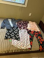 Set of six women's clothing items arranged on a bed: black and white houndstooth top, dark floral top, peach ruffled sleeveless blouse, black floral skirt, white cherry print sleeveless top, and navy blue floral pants.