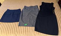 Three women’s office wear items: dark navy skirt, gray skirt, black dress on beige striped surface.