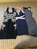 Lot of three women's party dresses laid on couch: black and cream embroidered dress, navy blue dress with pink straps, silver shimmer dress with tag.