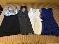 Four women's party dresses laid out on a bed, showing full length and styles including black/beige jeweled neckline, black polka dot halter, white lace peplum, and royal blue pleated