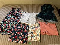 View of six clothing items laid on a couch including three tops and three bottoms: a black floral dress, white button-down top, dark green sleeveless top, and floral skirts and coral shorts.