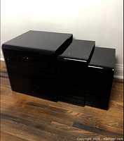 Set of three nested black glass tables shown from the front on a wood floor against a plain wall