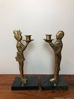 Pair of vintage brass candlesticks shaped as a man and woman standing on black marble bases, side view.