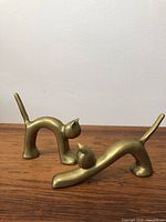 Two brass cat sculptures shown on wooden surface against white wall. Both cats have smooth stylized design, one stretching with elongated body, the other standing with arched back.