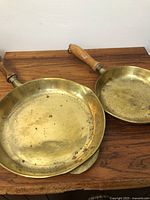 Two brass frying pans with wooden handles shown side by side, highlighting size difference and surface wear including spots of corrosion.
