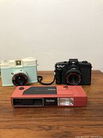 Three plastic cameras on wooden surface: cream/mint vintage camera, black Konica vintage camera, red Vivitar LF camera.