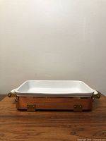 Front view of the casserole dish showing the white ceramic insert inside the copper caddy with brass hardware and white handles.