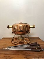 Full view of the copper fondue pot on its stand alongside eight fondue forks.