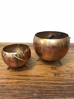 Two copper plant pots on wooden surface, one smaller with handle and one larger without handle, both on three small legs, showing some wear and tarnish