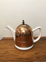 Front view of the vintage tea pot showing white ceramic handle and spout with copper insulated hood topped by brass finial.