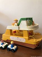 Side view of the Fisher Price ferry boat toy with three figures seated on the upper deck and two small cars placed in front of the boat on the floor.