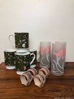 Four vintage ceramic mugs with green and pink floral patterns stacked on a wood surface along with three frosted glass tumblers and four hand-painted ceramic napkin rings.
